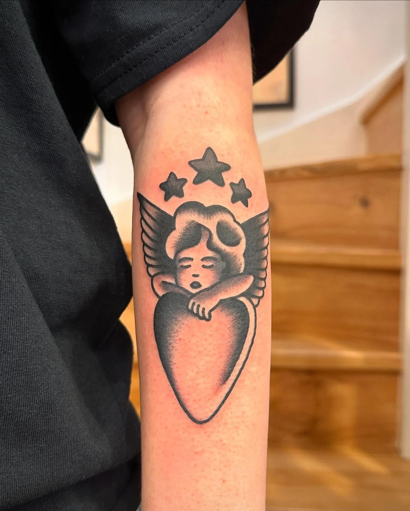 Tattoo of an angelic figure with closed eyes, curly hair, and wings, hugging a large heart, with three stars above the head.