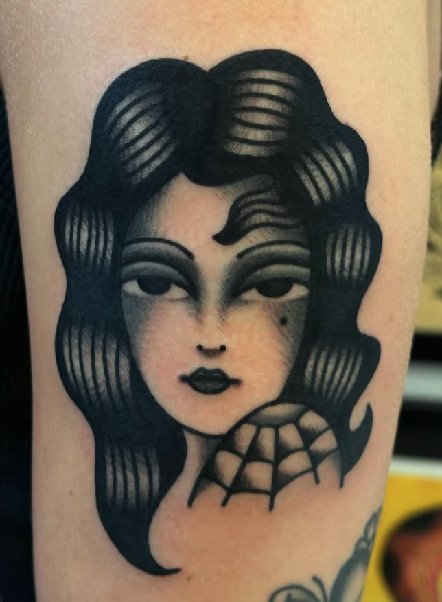 Black and gray tattoo of a woman with long hair and bold makeup, with a spiderweb design below her chin.