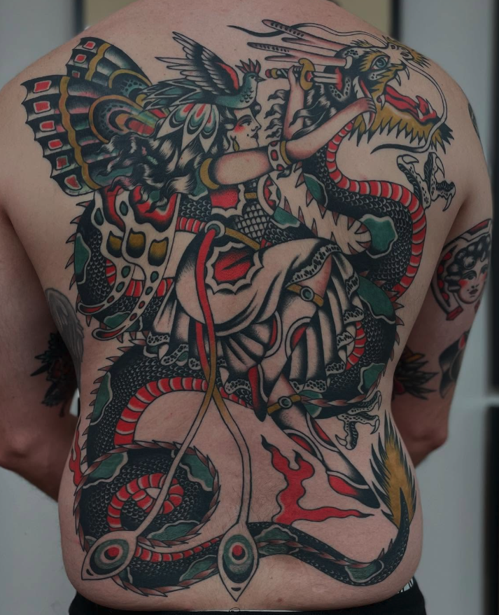 A tattoo of a woman in fantasy armor holding a spear, riding a dragon. The tattoo covers the chest and stomach, with intricate details and bold colors including black, red, yellow, and green.