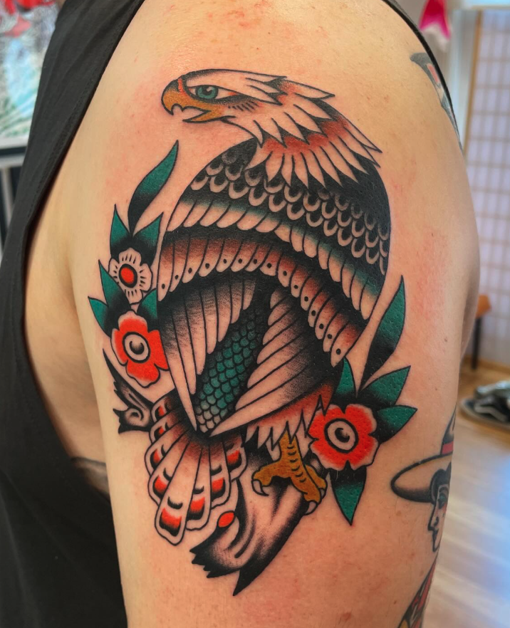Traditional style tattoo of an eagle's head and body perched on a branch with orange and white flowers on the upper arm.