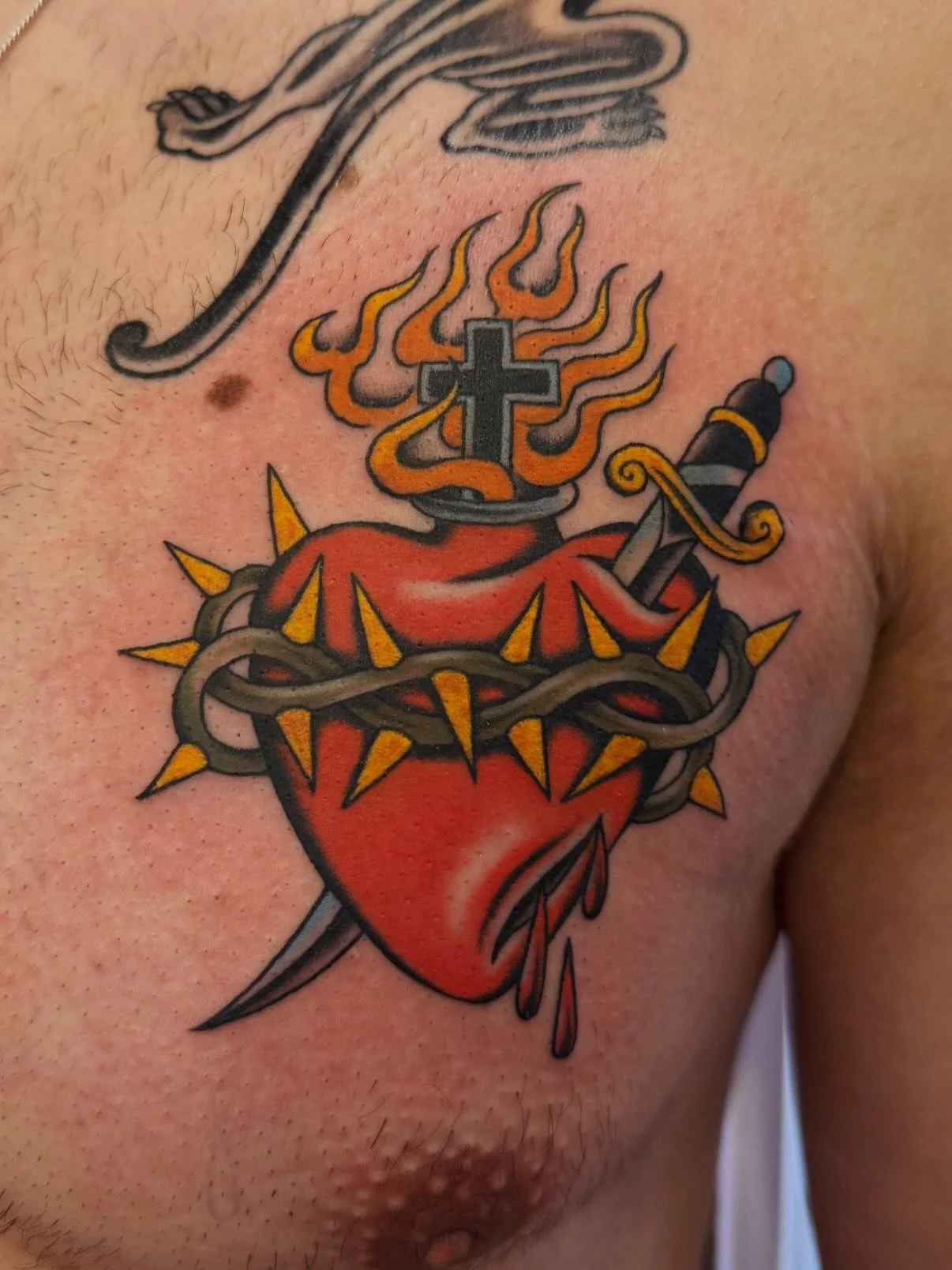 Tattoo of a realistic red human heart with a black cross on top, surrounded by flames, with a sword piercing through the heart and a crown of thorns around it.