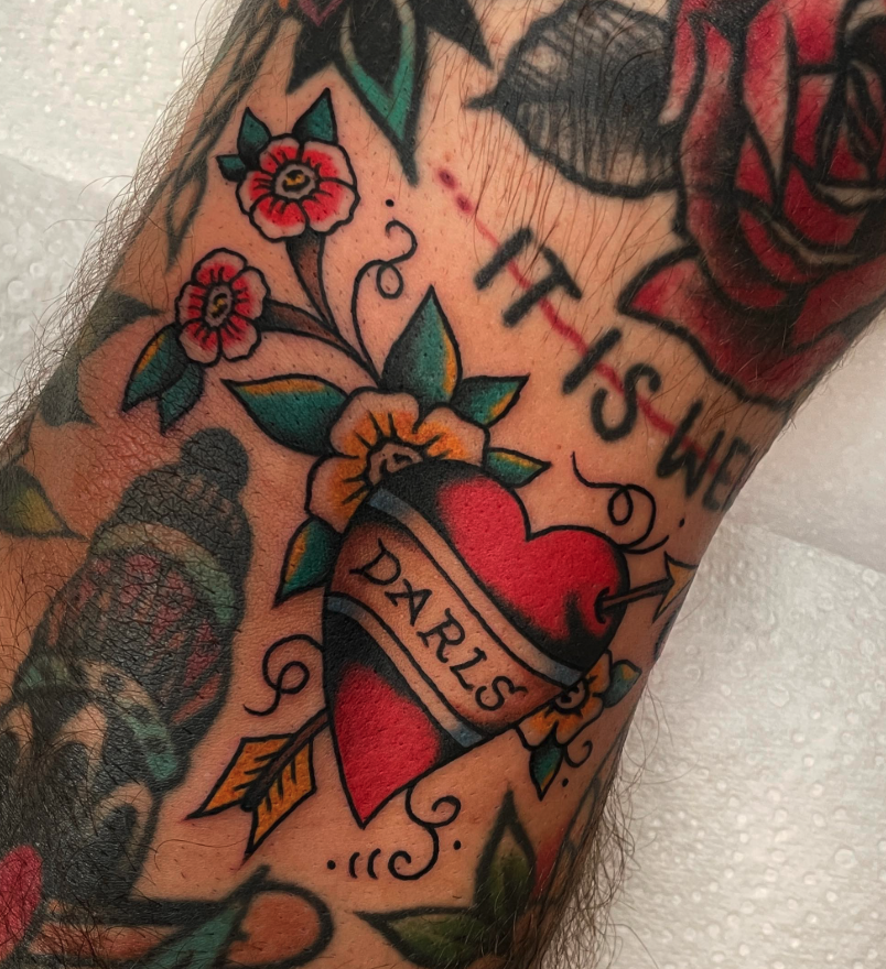 A tattoo of a red, anatomically correct heart with a banner reading 'Daughters' wrapped around it, surrounded by flowers and leaves.