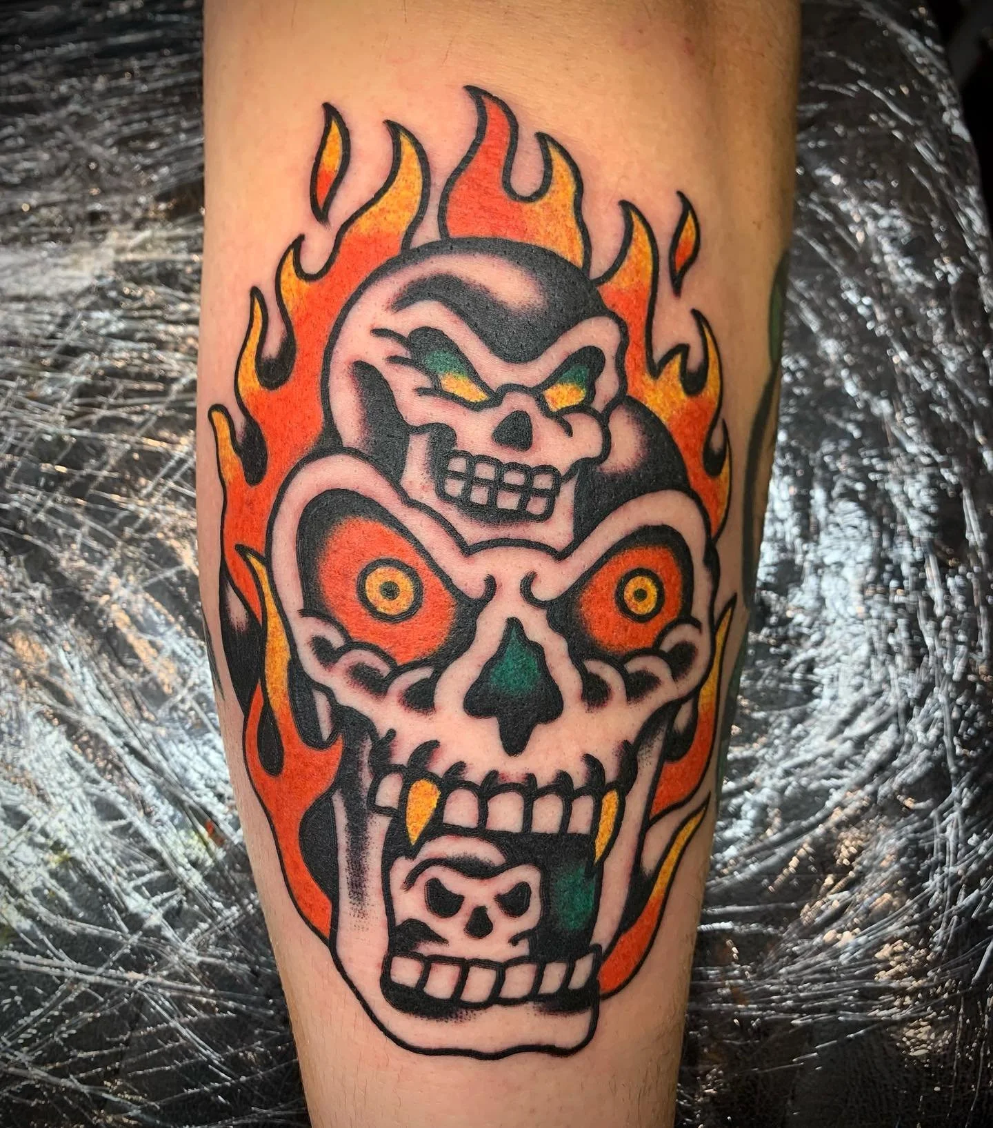Tattoo of a flaming skull with exaggerated features, bright colors, and bold outlines, resembling traditional tattoo style.