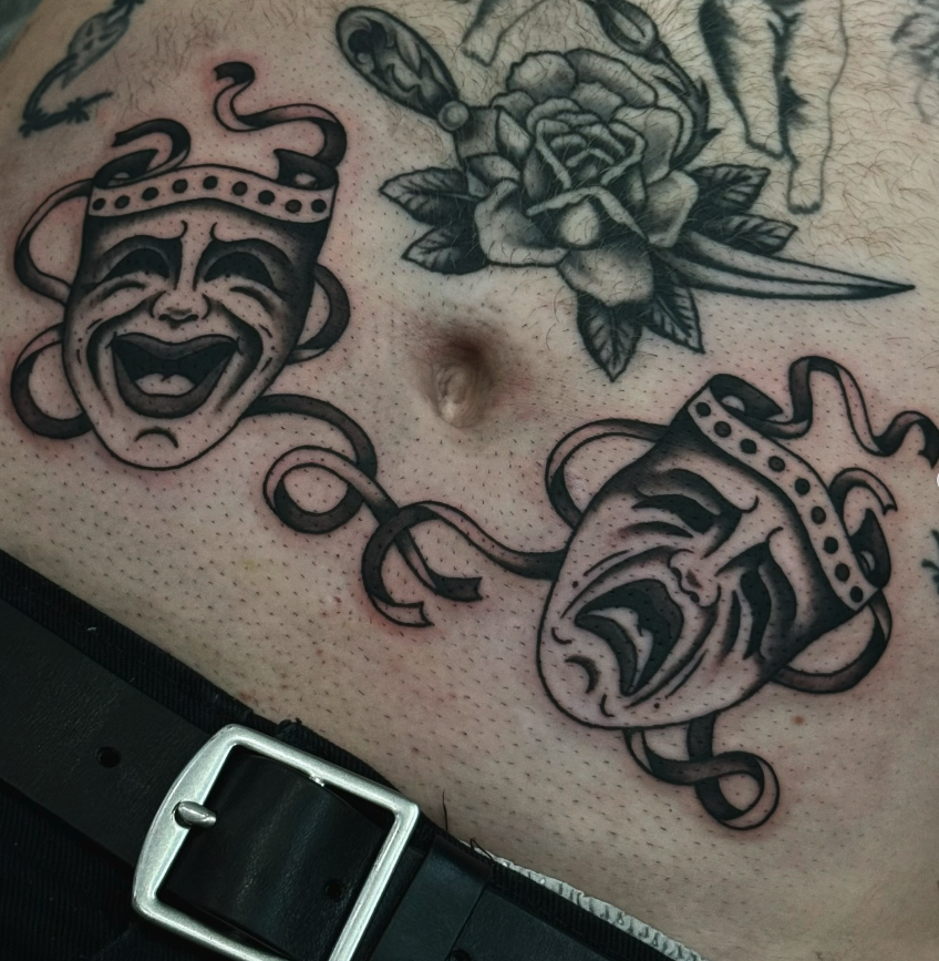 Tattoo of two theatrical masks, one smiling and one crying, connected by a ribbon, with a detailed black and gray rose above them.