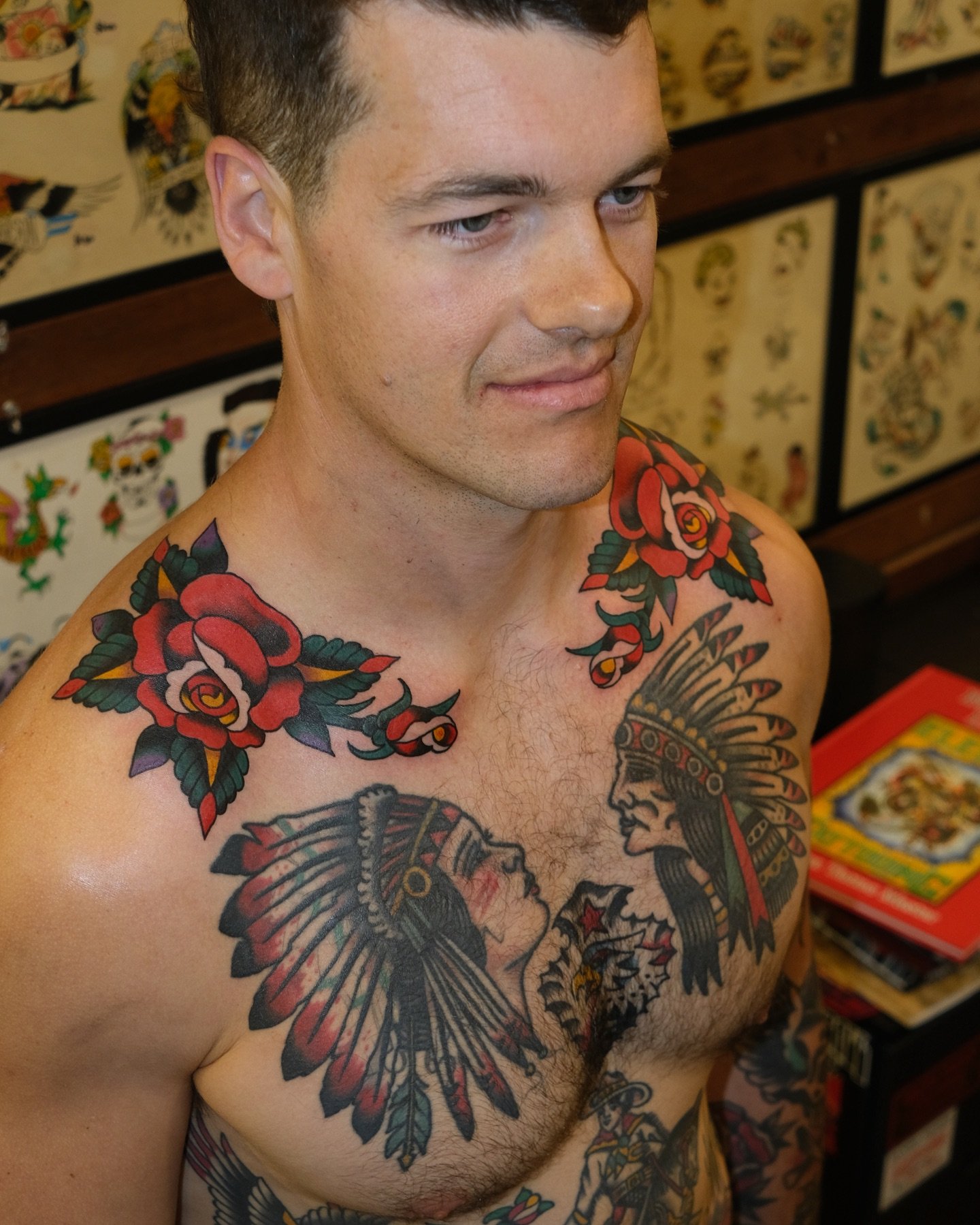 A shirtless man with tattoos of roses on his shoulders and Native American chief portraits on his chest, standing in a room with tattoo designs on the wall.