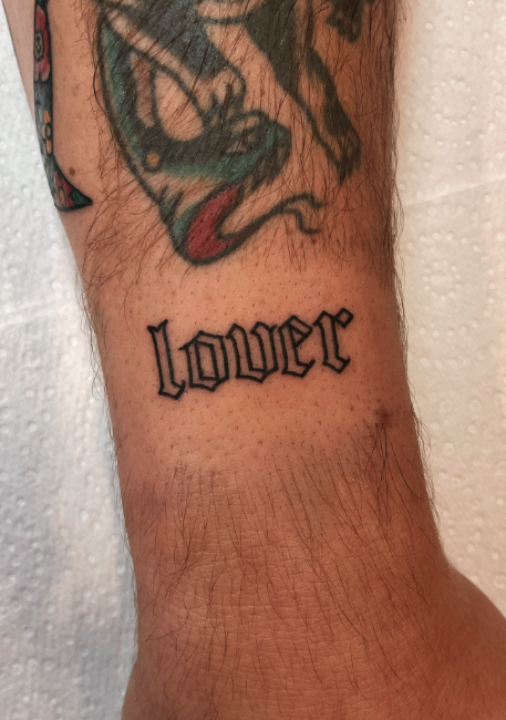 Tattoo of the word 'Lover' in black Gothic script on a person's forearm, with part of a colorful tattoo above.