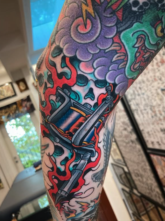 Colorful tattoo on arm depicting a tattoo machine surrounded by smoke, clouds, and stylized flames with vibrant colors.