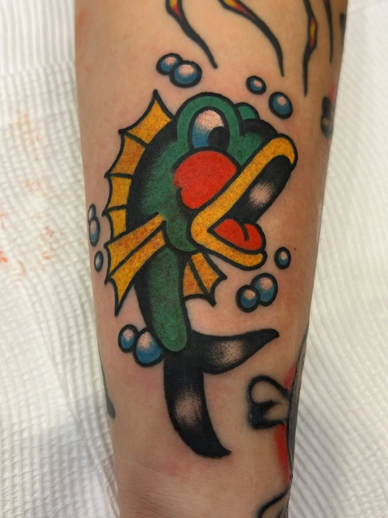 Traditional style tattoo of a cartoonish, colorful fish with a red mouth, blue eyes, yellow fins, and bubbles around it.