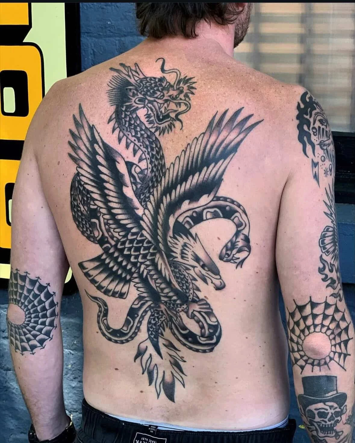 A man with large black tattoos on his back and arms, including a dragon, a snake, spider webs, and other designs.