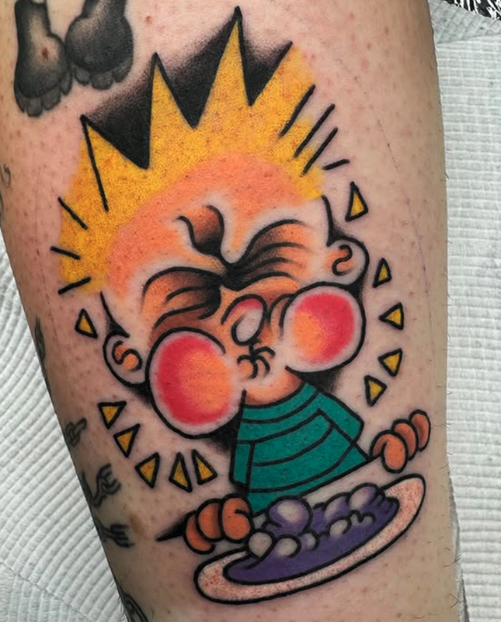 Tattoo of a cartoon child with rosy cheeks, orange hair, wearing a teal striped shirt, sitting on a plate of food with grapes, under a yellow sun with triangular rays.
