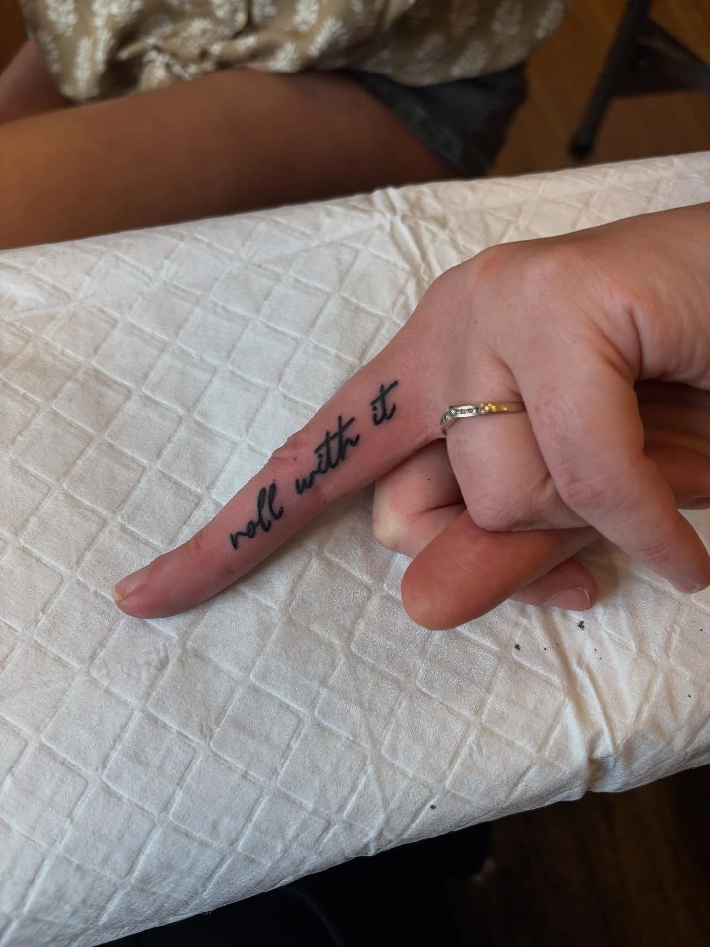 A hand with a tattoo on the index finger that reads 'not with it' in black ink. The person is wearing a thin ring on the ring finger. The hand is resting on a quilted white surface.