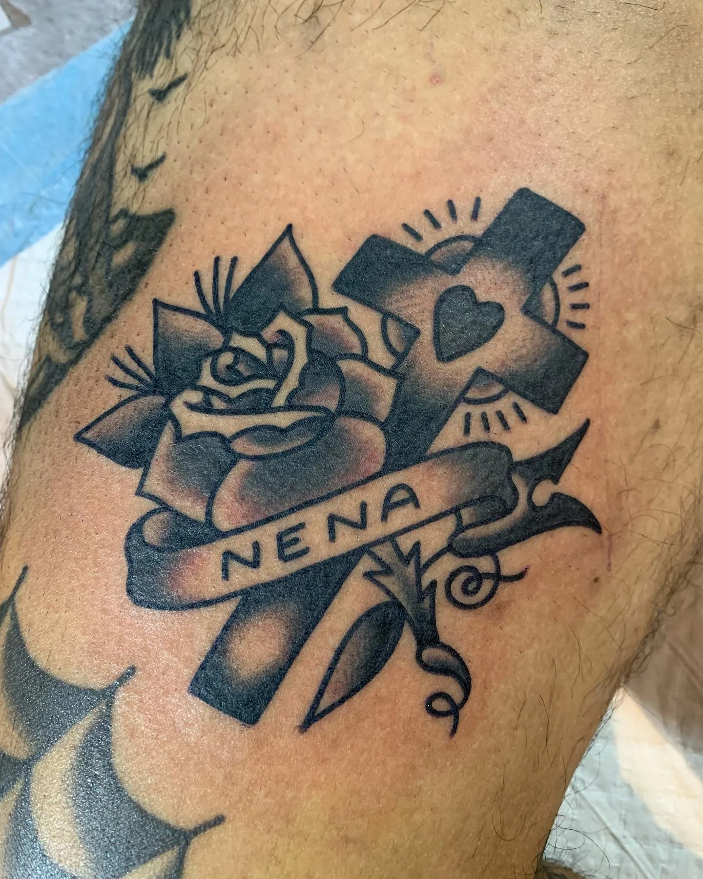 Tattoo of a rose flower with the word 'NENA' on a banner, a cross with a heart in the center, and decorative elements.