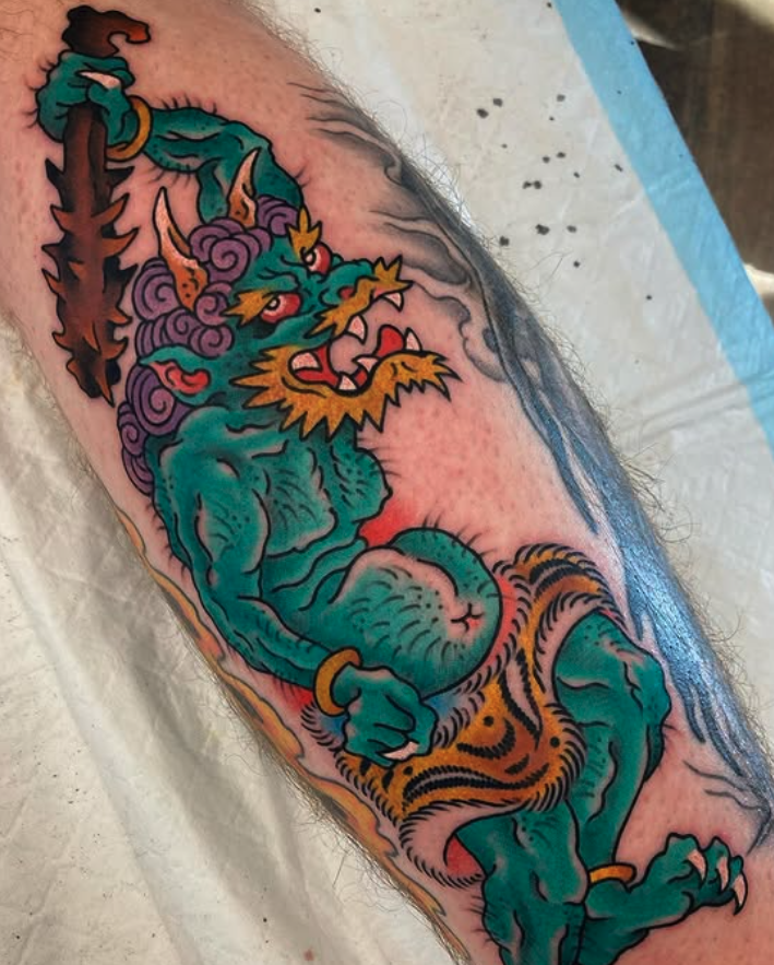 Colorful tattoo of a fierce dragon with green scales, yellow accents, purple flames, and red eyes on a person's arm.