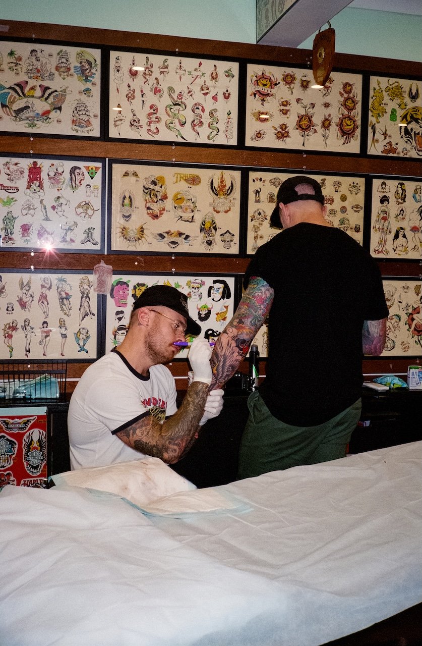 Two tattoo artists, one seated and one standing, working on tattoos. The seated artist is tattooing the standing artist's arm. The background features framed tattoo flash art on the wall.
