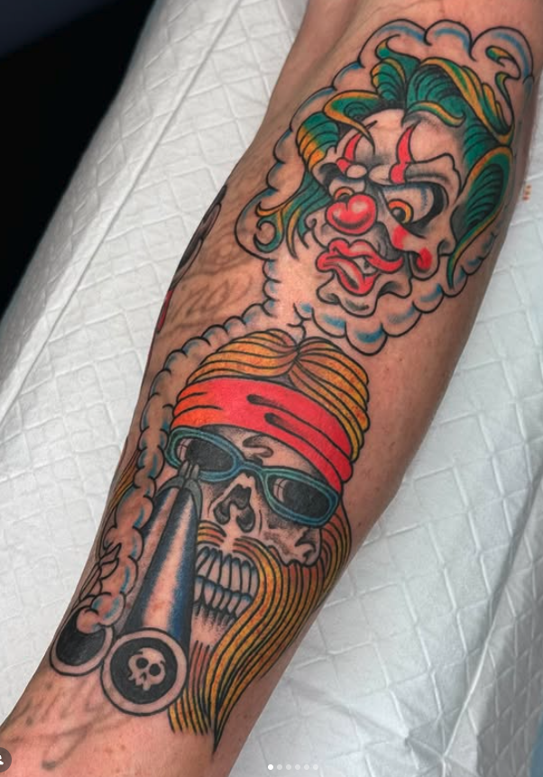 Tattoo of a skull with sunglasses and a beard, smoking a bong, accompanied by a cartoonish, colorful clown face with green hair and a red nose in the cloud above the skull.