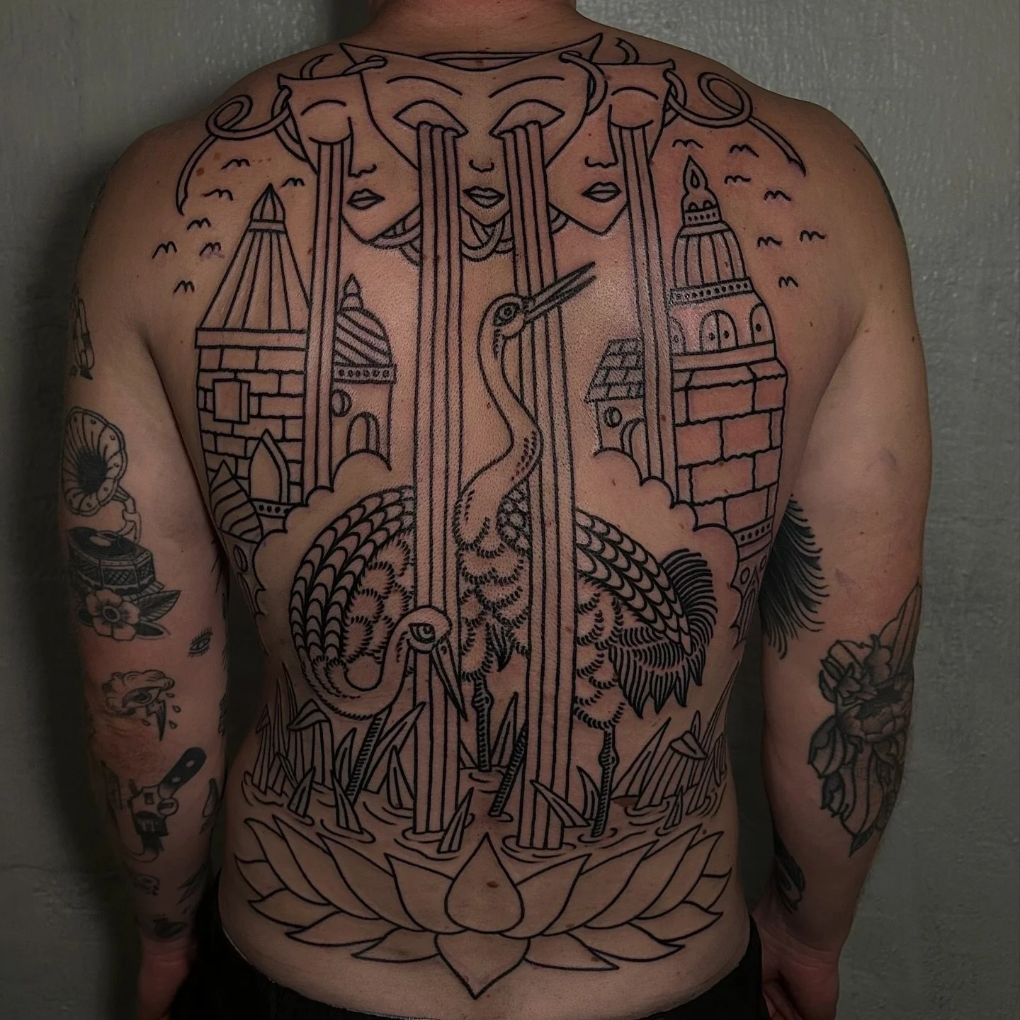A large tattoo covering the person's chest and abdomen featuring a stylized design with multiple faces, a crane, buildings, and a lotus flower at the bottom, all in black ink.