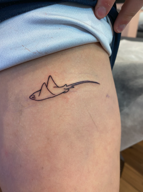 Tattoo of a minimalist shark outline on person's thigh.