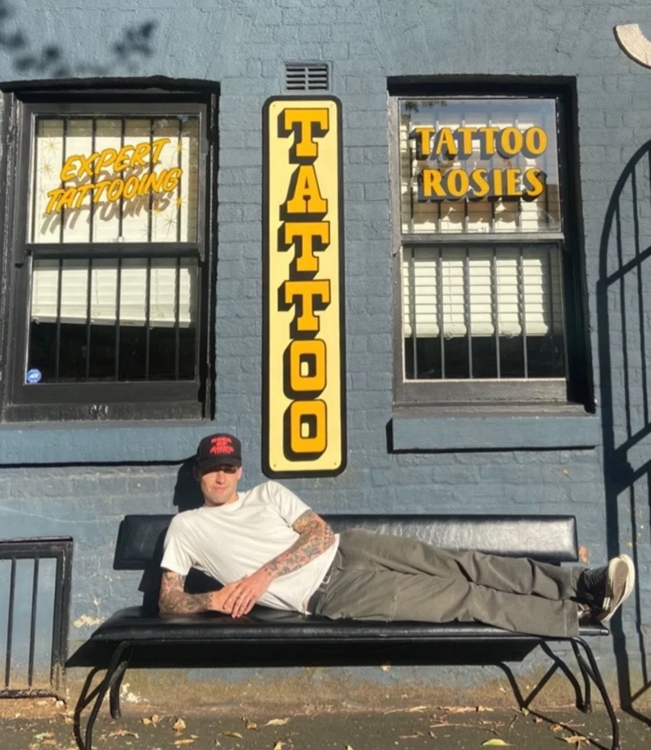 A man lying on a black bench in front of a blue brick building with windows that have yellow text advertising a tattoo shop. The left window says "EXPERT TATTOOING," and the right window says "TATTOO ROSIES." A vertical yellow and black sign in the center reads "TATTOO." The man is wearing a white t-shirt, gray pants, black sneakers, a black cap, and has tattoos on his arms.