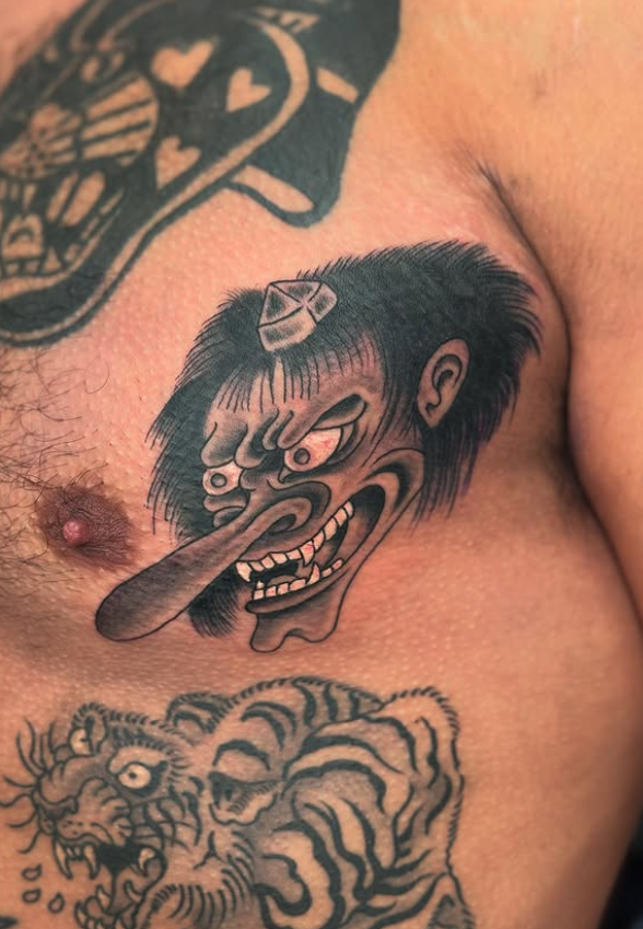 Tattoo of a monstrous face resembling a creature with exaggerated features, a long tongue, and a wild mane; part of a larger tattoo of a roaring tiger below, on a person's chest.
