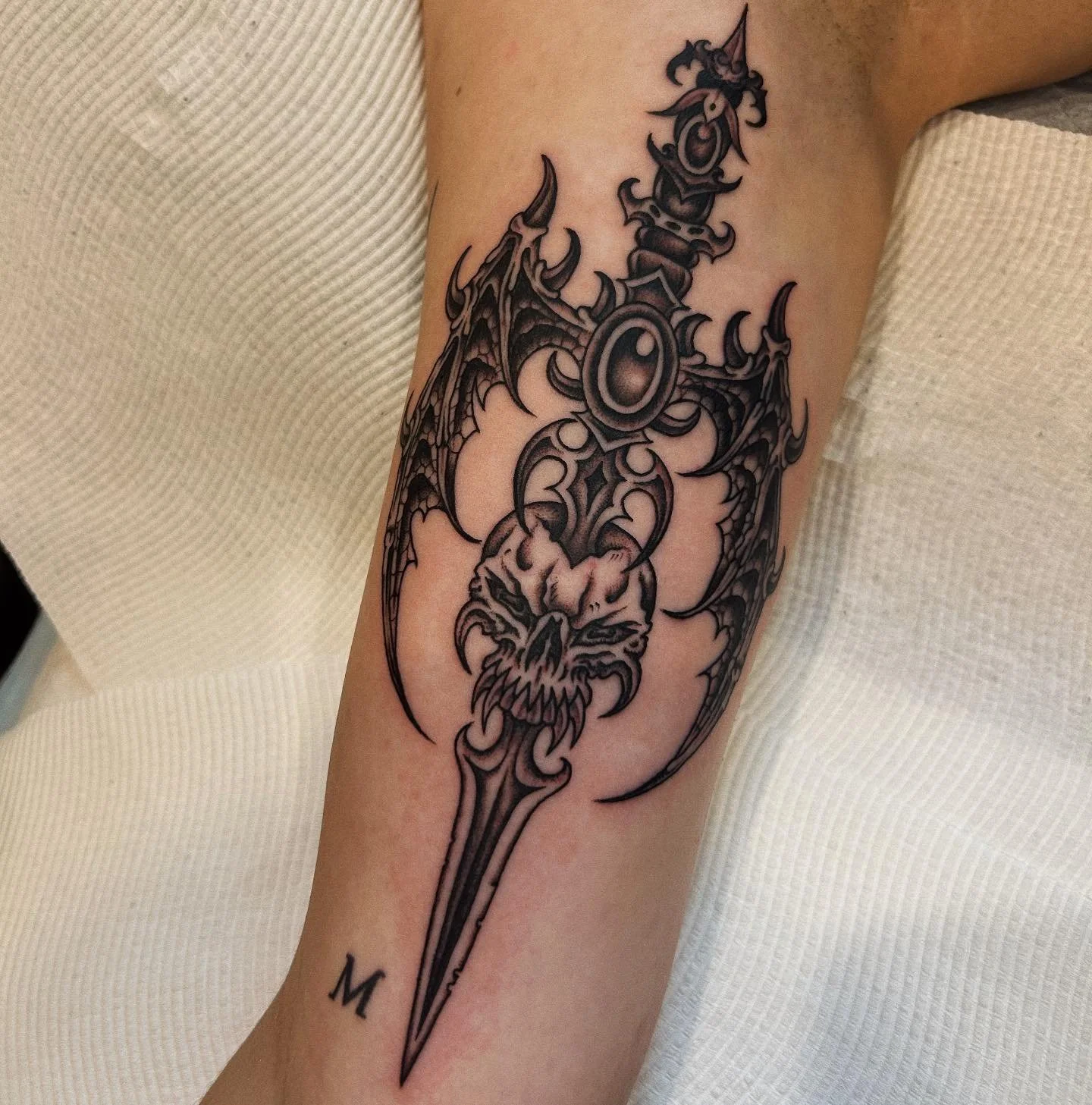 Black and gray tattoo of a demonic sword with bat-like wings and a fierce skull face on the forearm of a person.