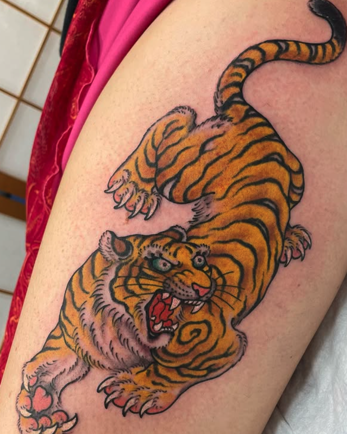 Colorful tattoo of a fierce tiger with orange and black stripes, showing sharp teeth and blue eyes, positioned on a person's arm.