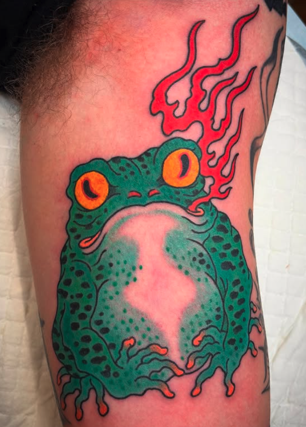 Tattoo of a colorful frog with orange eyes and red flames emerging from its head, on a person's upper arm.