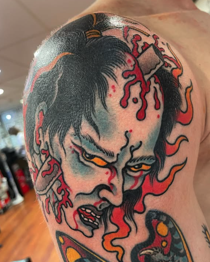 Close-up of a tattoo on a person's upper arm and shoulder featuring a stylized, fierce face with black hair, intense yellow eyes, and red flames or energy emanating from the face. The artwork has bold black outlines and vibrant colors.