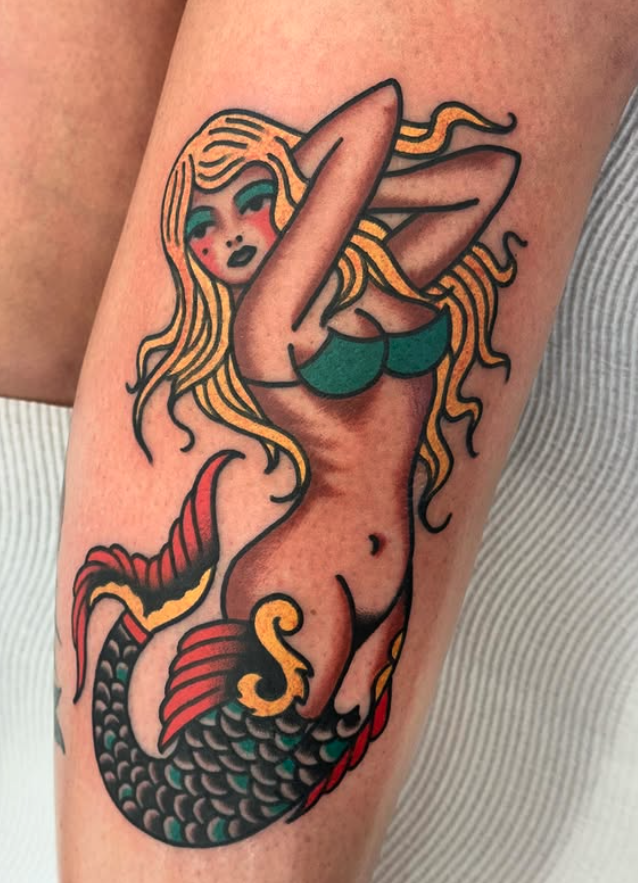 Tattoo of a mermaid with blonde hair, green top, and a colorful tail in red, black, and green, on a person's arm.