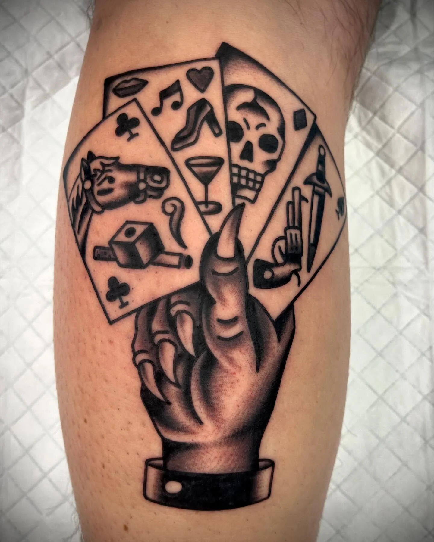 A tattoo of a hand with a nail, holding a circular object divided into six sections, each containing different symbols: a music note, a skull, a syringe, a knife, a whiskey glass, and a clown face, arranged in a semi-circle.