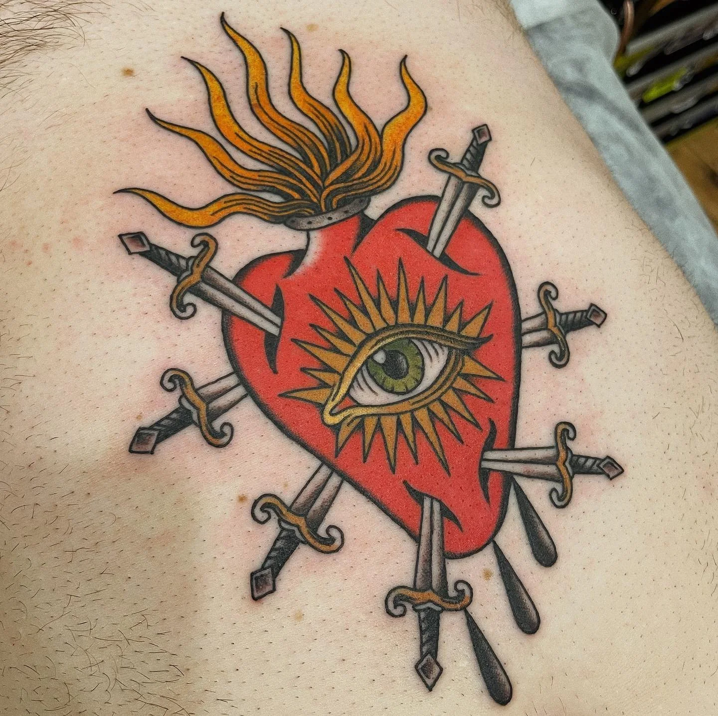A tattoo of a red heart pierced by multiple swords, with a large eye in the center surrounded by a sunburst design, and flames coming out of the top of the heart.
