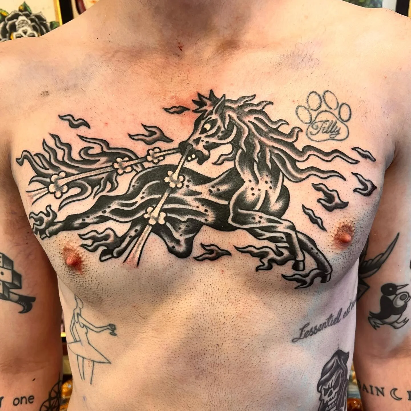 A tattoo of a black and gray fierce unicorn with flames on its mane and tail, rearing up on its hind legs, holding a flaming spear in its mouth, on a person's chest.