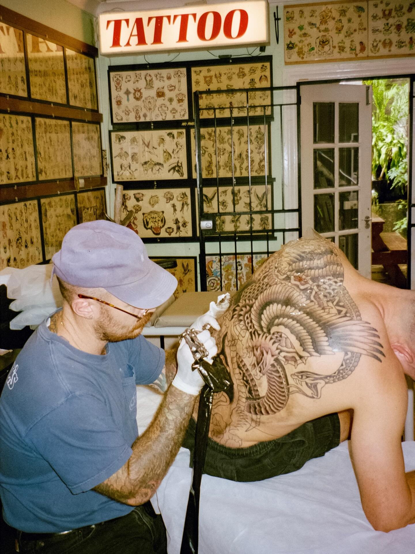 Tattoo artist in a gray cap and glasses tattooing a large dragon design on a man's back in a tattoo studio with tattoo flash images on the walls and a sign that says 'TATTOO'.