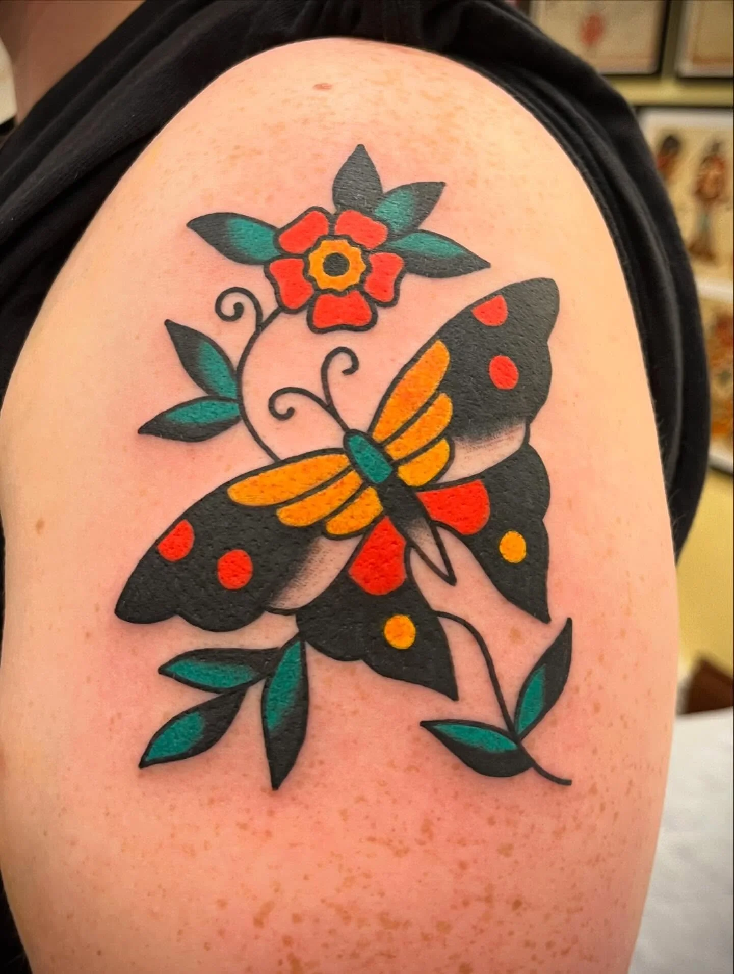 A colorful tattoo of a butterfly with black wings, yellow and orange details, and red spots, surrounded by a flower with red petals and green leaves on a person's upper arm.