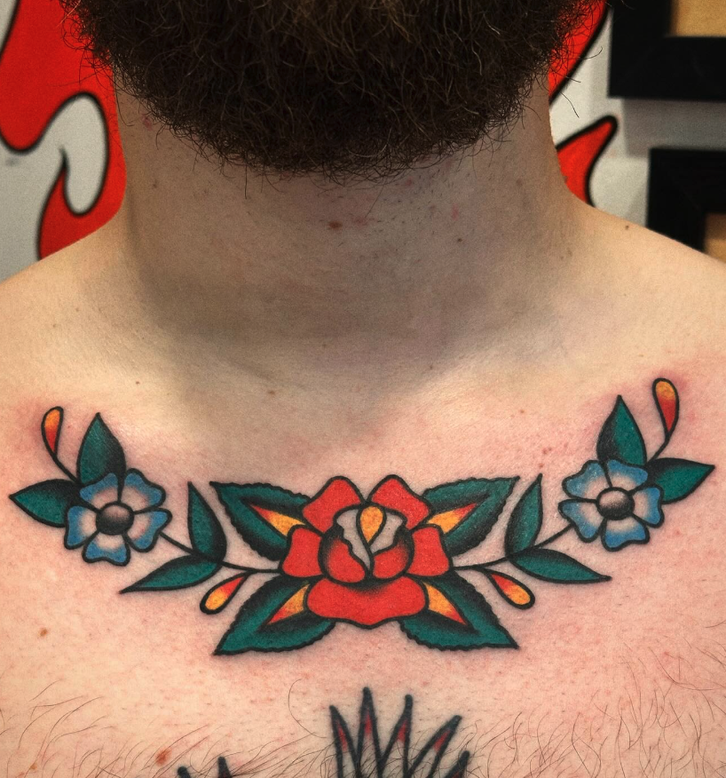 Colorful tattoo of a central red rose with green leaves and blue flowers on each side on a person's upper chest.