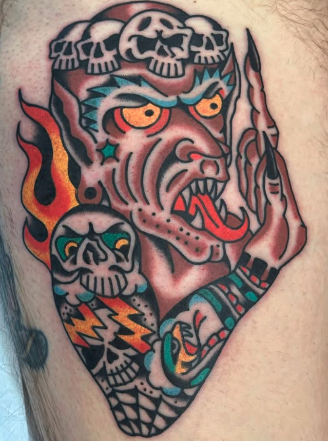 Tattoo of a demonic face with skulls on its head, flames, a skull with lightning bolts, and tattooed arm with tribal patterns.