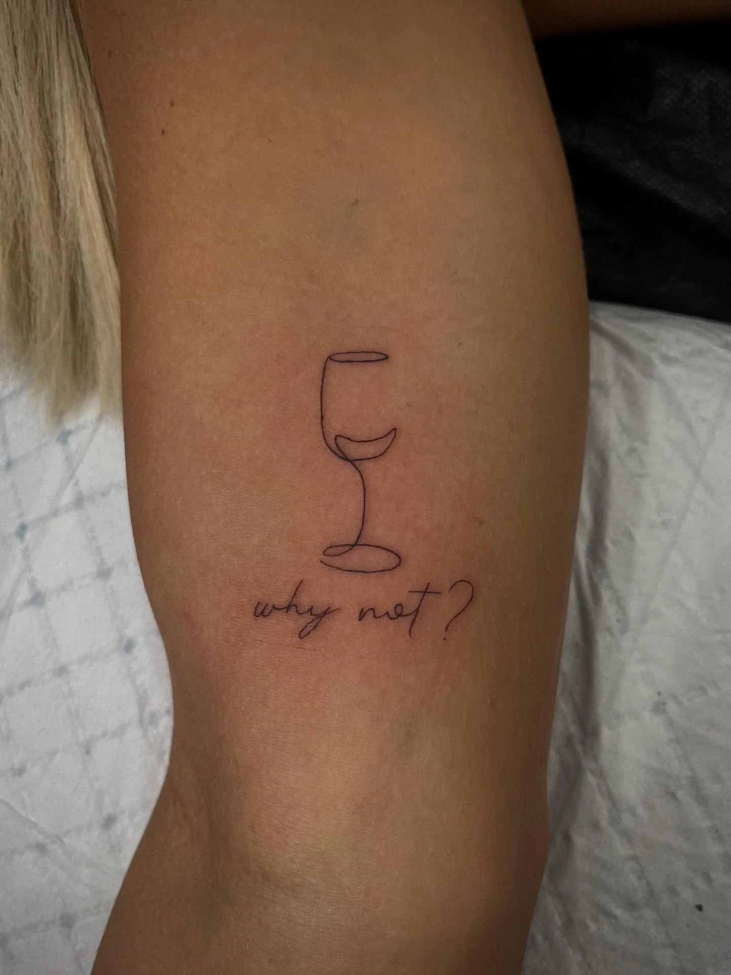 Tattoo of a wine glass outline with the phrase 'why not?' written below on a person's arm.