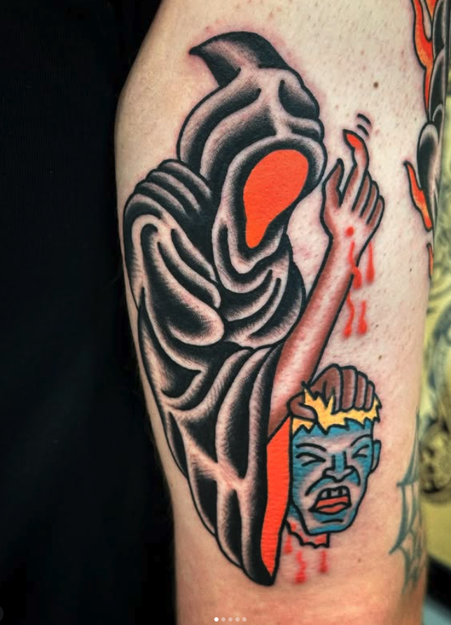 Tattoo of a stylized, dark figure with a face-shaped orange area, holding a small, colorful, distressed face with blue skin and yellow hair, dripping with red ink.