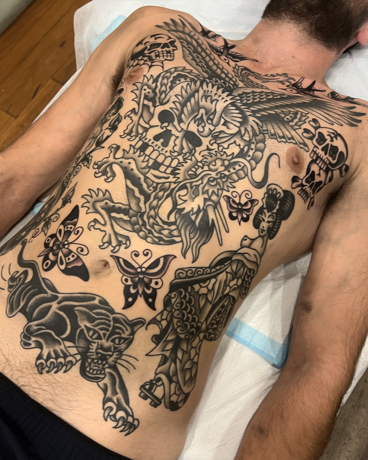 A person's chest with various black ink tattoos, including a skull, a dragon, butterflies, a panther, and a woman, in a traditional tattoo style.