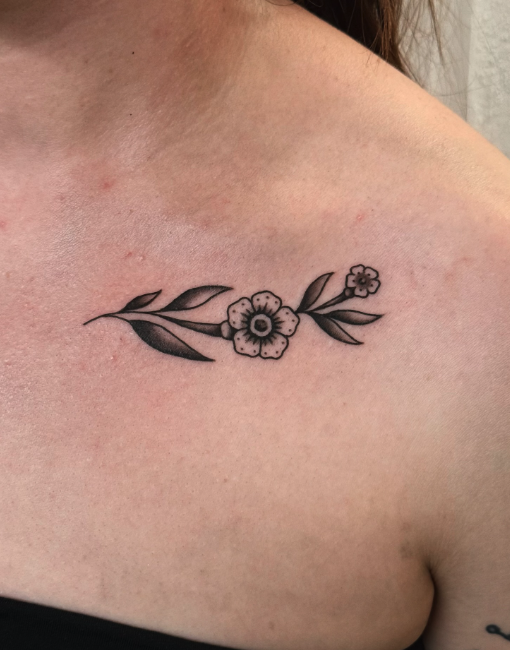 Black and gray floral tattoo on person's collarbone featuring a central flower with leaves extending outward.