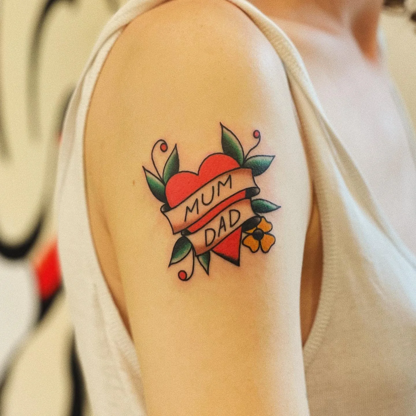 Tattoo of a red heart with green leaves and yellow flowers, with a banner reading 'MUM DAD' on an upper arm.