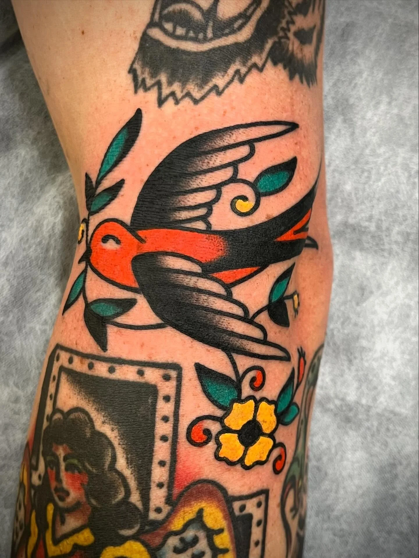 Colorful tattoo of a flying bird with orange and black feathers, holding a small leaf in its beak, surrounded by leaves, flowers, and framed portraits.