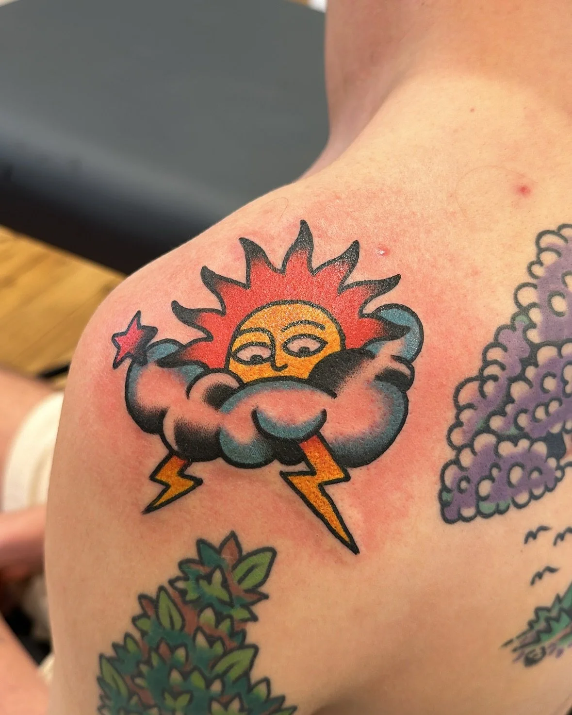 Tattoo of a smiling sun with closed eyes, surrounded by clouds and lightning bolts on a person's shoulder.