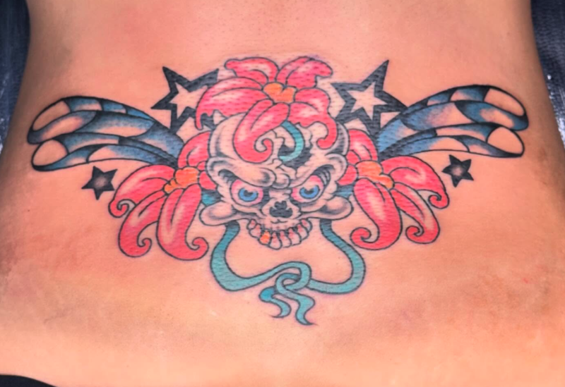 Tattoo of a skull with flowing pink hair, blue eyes, and a snarl, with black wing-like shapes and stars behind it on skin.