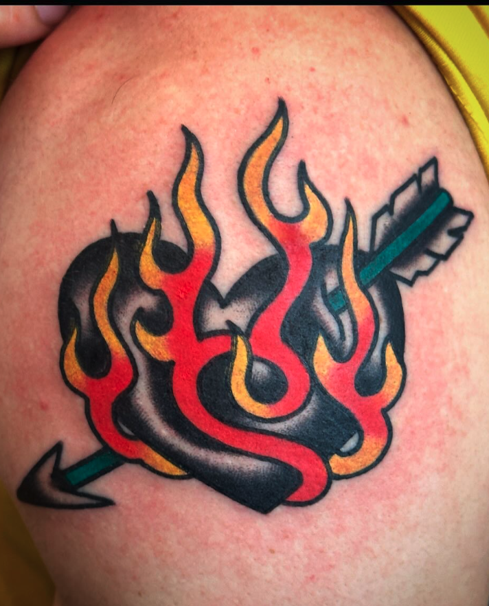 Tattoo of a flaming heart pierced by an arrow, with flames in red, orange, and yellow, on a person's skin.