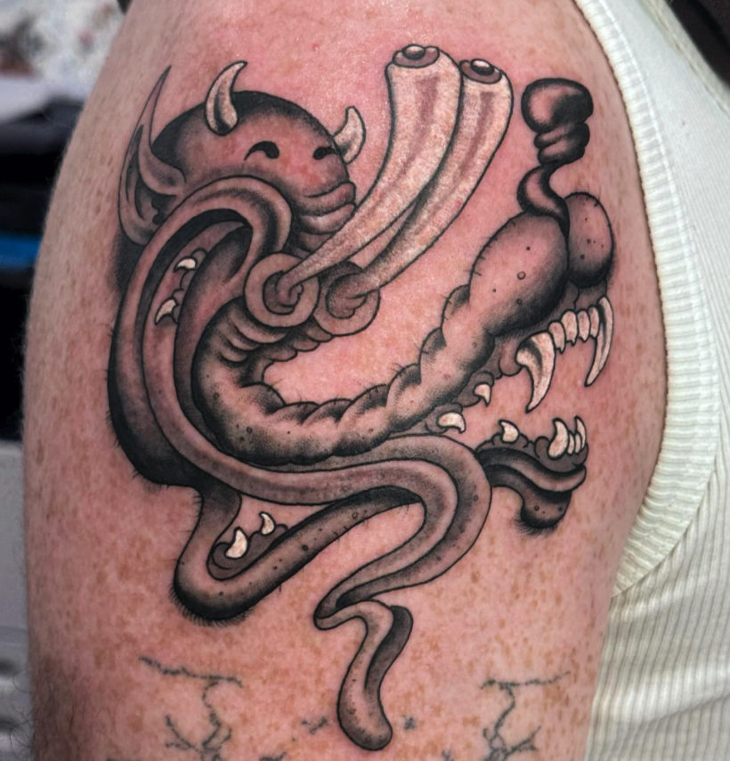 Tattoo of a cartoonish dragon with a content expression, large eyes, horns, and exaggerated features, depicted in black and grey ink.