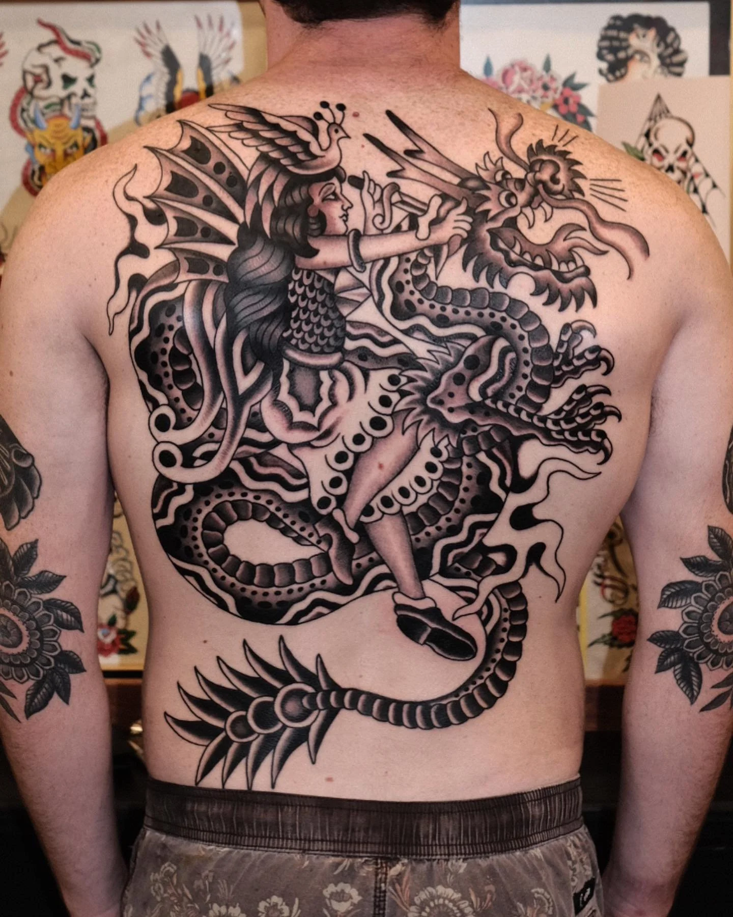 Black and gray tattoo of a woman with butterfly-like wings riding a dragon, covering the back. The woman has long flowing hair and is in a dynamic pose.