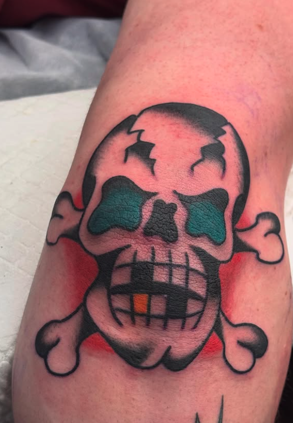 Tattoo of a skull and crossbones with a cartoonish style, featuring green eye sockets, a cracked top, and a mouth with orange and black teeth.