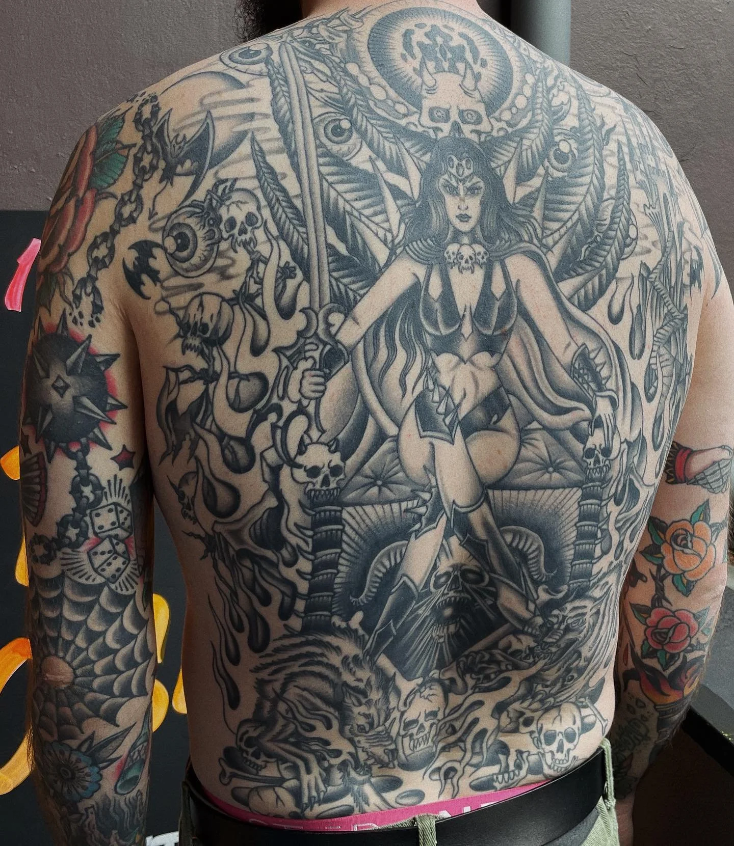 Black and white tattoo of a female warrior with a crown, crowned skulls, and weapons, surrounded by various skulls, flames, dice, and floral elements on a person's back and arms.