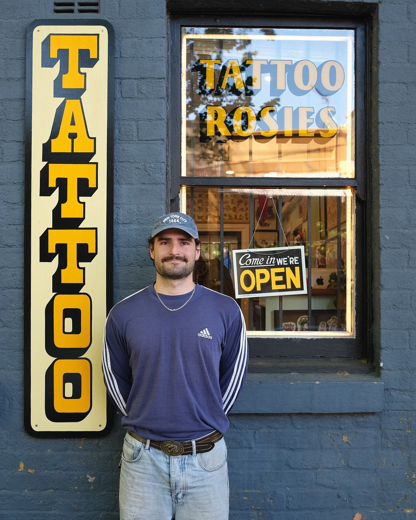 A man standing in front of a tattoo shop with signs reading "TATTOO" and "TATTOO ROSIES" outside a blue brick building, with an "OPEN" sign hanging in the window.