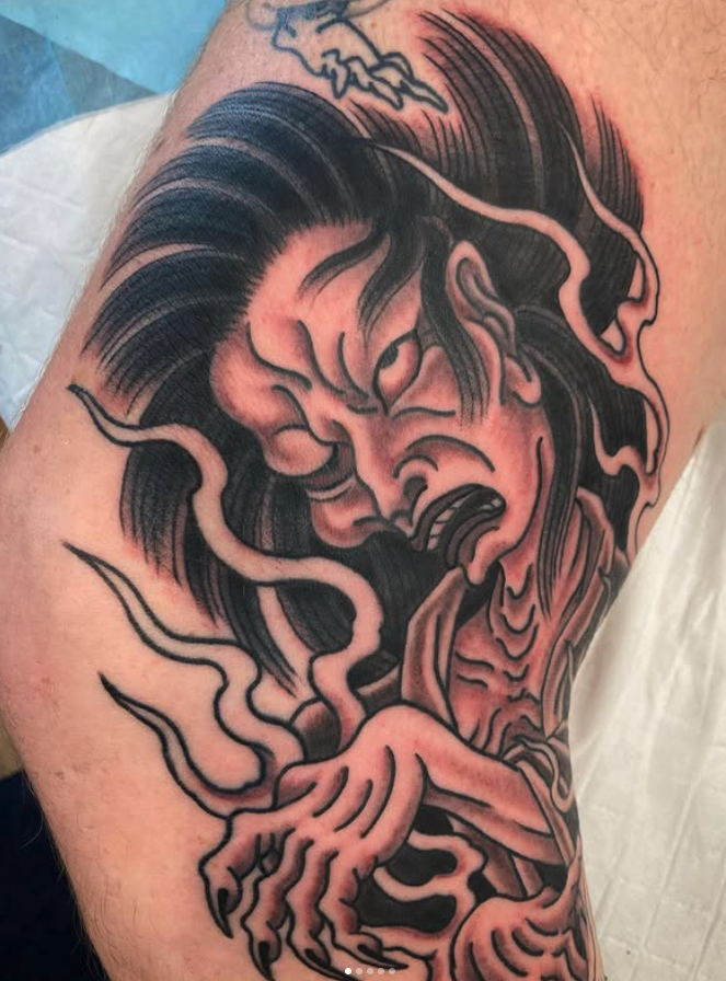 Tattoo of a fierce, stylized demon or oni with sharp claws, flowing hair, and smoke-like elements in black and gray ink on arm.